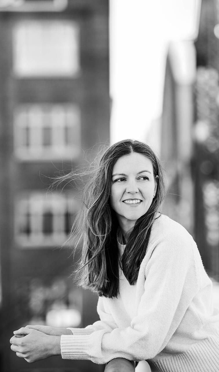 A black and white photo of the yoga instructor standing on a bridge in Portland, Oregon. The person is smiling and gazing away from the camera, with the wind gently tousling their hair. This image captures a moment of tranquility and connection with nature